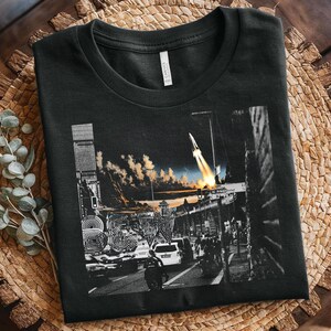 Könnte beinhalten: Schwarzes T-Shirt mit einem Graustufen-Grafik einer Stadtstraßenszene mit einem Raketenstart. Das Design umfasst Gebäude, Fahrzeuge und Menschen mit einem lebendigen Himmel. Das Shirt besteht aus einem weichen Material.