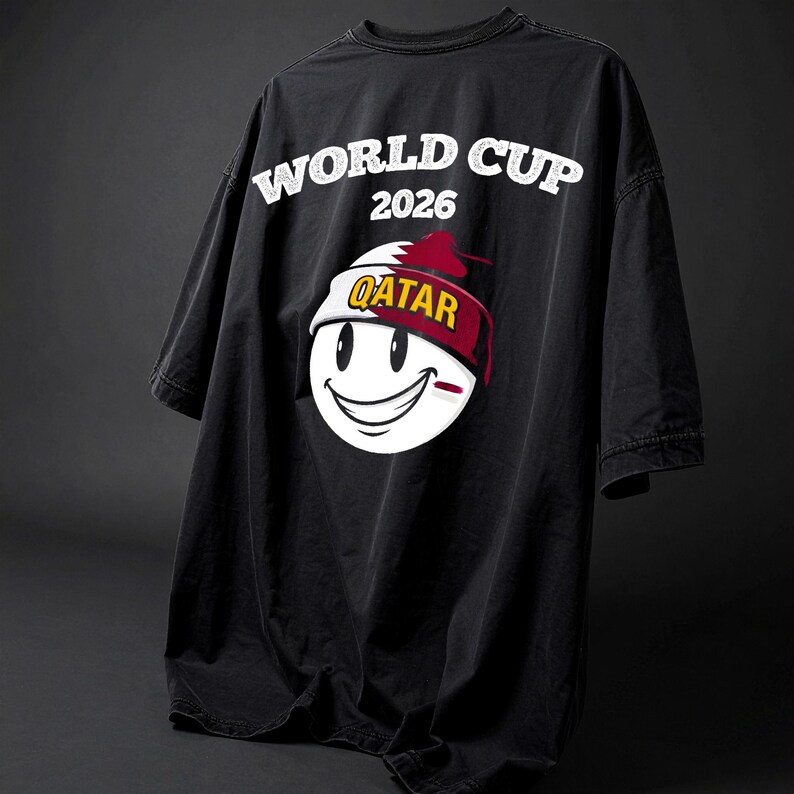 May include: Black oversized t-shirt with the text "WORLD CUP 2026" in white. The shirt features a smiling face graphic wearing a hat with the word "QATAR" on it. The hat is red and white.