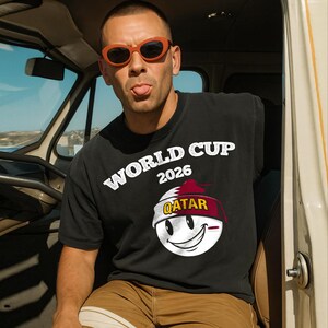 May include: Black t-shirt with the text "WORLD CUP 2026" in white. Below the text is a graphic of a smiling face with the Qatar flag colors. The person in the image is wearing orange sunglasses.