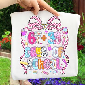 May include: White t-shirt with a colorful graphic celebrating "67 + 33 Days of School." The design includes a pink and white checkered bow, school supplies, and a floral pattern. The shirt is held up by a hand against a background of green foliage and flowers.