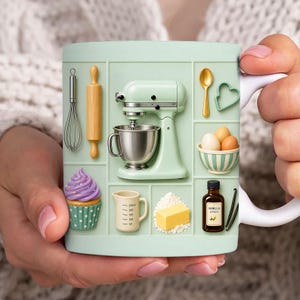 May include: A mint green mug with baking-themed illustrations. The design includes a stand mixer, whisk, rolling pin, measuring jug, butter, eggs, a cupcake, and a vanilla extract bottle. The mug is held by a person.