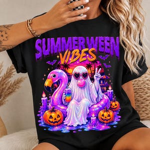 Puede incluir: Una camiseta negra con un estampado gráfico con el texto "SUMMERWEEN VIBES" en morado y naranja. El diseño muestra un fantasma blanco con gafas de sol en forma de corazón, haciendo una burbuja rosa, sentado en un flotador de flamenco rosa con calabazas.