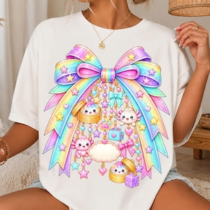 May include: A white t-shirt featuring a large, colorful rainbow-gradient bow graphic. The bow is adorned with dangling charms including small axolotls, dim sum in bamboo steamers, stars, hearts, a small blue suitcase, and a purple cube.