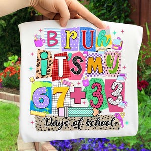 May include: White t-shirt with a colorful graphic design featuring the words "BRU ITS MY 67+33 Days of school." The design includes various school-themed elements like pencils, scissors, and a backpack.