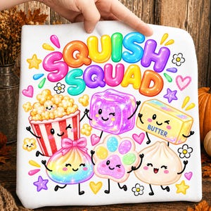 Può includere: Una t-shirt bianca con il testo colorato SQUISH SQUAD sopra illustrazioni a cartoni animati di un secchiello di popcorn, un cubo viola, un panetto di burro, un raviolo arcobaleno, un'impronta di zampa e un raviolo bianco, tutti sorridenti.