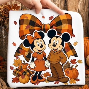 May include: A white sweatshirt featuring a graphic of Minnie and Mickey Mouse dressed in autumn attire. They stand before a large orange and brown plaid bow, surrounded by pumpkins and scattered fall leaves in shades of orange, red, and yellow.