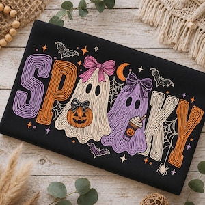 May include: A black t-shirt featuring the word SPOOKY in large, colorful, textured lettering. The design includes two ghosts wearing bows, one holding a jack-o'-lantern and the other a coffee cup, surrounded by bats, a spider, and a crescent moon.