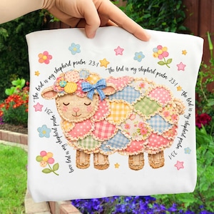 May include: White tote bag with a patchwork sheep design, featuring a blue bow and floral details. The text "the lord is my shepherd psalm 23:1" is printed around the sheep. The bag is held by a hand.