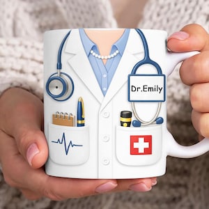 Puede incluir: Taza de cerámica blanca con diseño de bata de médico, que incluye un estetoscopio, un bolígrafo y suministros médicos. La taza tiene el nombre "Dr. Emily" y un símbolo de cruz roja.
