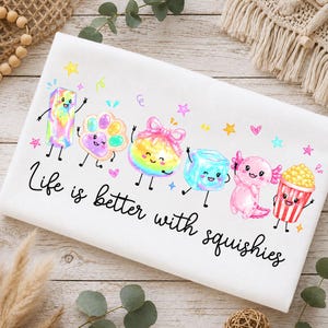 May include: A white t-shirt featuring a colorful graphic of six cartoon characters including a rainbow block, a paw print, a round pouch, an ice cube, an axolotl, and popcorn, with the text Life is better with squishies in black script.