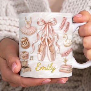 May include: White ceramic mug featuring ballet-themed illustrations. The design includes pink ballet shoes, a tutu, a swan, and the name "Emily" in gold script. Other elements include a music note, a jewelry box, and a miniature record player.