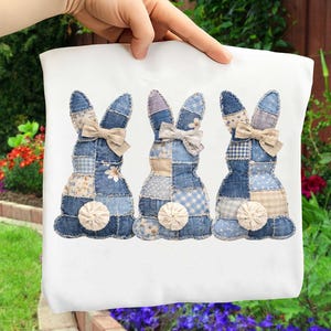May include: White tote bag with three denim patchwork bunny designs. Each bunny has a beige bow and a fluffy white tail. The bunnies are made from various blue patterned fabrics, including floral and gingham patterns.