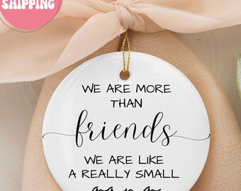May include: A white ceramic ornament with the text "WE ARE MORE THAN friends WE ARE LIKE A REALLY SMALL gang" in black script. A small pink heart is at the bottom. The ornament hangs from a gold string.