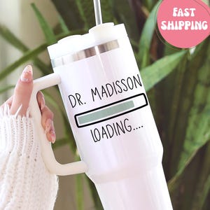 May include: White insulated tumbler with a handle and a straw. The tumbler features the text "DR. MADISSON LOADING...." with a loading bar graphic. A pink circle with the words "FAST SHIPPING" is visible.