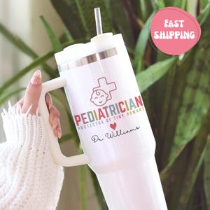 May include: White insulated tumbler with a handle and straw. The tumbler features the word "Pediatrician" in red and the phrase "Protector of Tiny Humans." A red heart and a cross with a baby face are also visible. The name "Dr. Williams" is printed below.