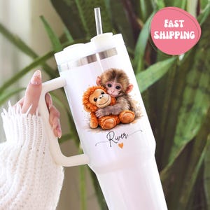 May include: White tumbler with a handle, featuring a watercolor illustration of a baby monkey hugging a stuffed animal monkey. The name "River" is written below the image, with a small orange heart. Includes a white lid and straw.