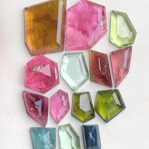 Peut inclure: Une collection de pierres précieuses à facettes de différentes couleurs, dont le rose, le vert, le jaune et le bleu. Les pierres précieuses sont taillées en formes géométriques, telles que des hexagones et des rectangles. Les pierres précieuses sont disposées sur une surface blanche.