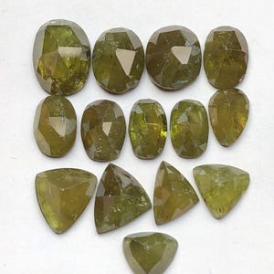 Könnte beinhalten: Eine Ansammlung facettierter, olivgrüner Edelsteine in verschiedenen Formen, darunter oval, kissenförmig und dreieckig. Die Edelsteine sind auf einer weißen Oberfläche angeordnet, um ihre Klarheit und Farbe zu zeigen.