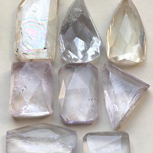 May include: A collection of faceted gemstones in diverse shapes, such as teardrop, rectangular, and irregular forms. The crystals are mainly clear with delicate shades of lavender and yellow. The gemstones are displayed on a neutral background.