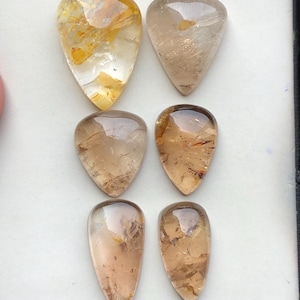 以下が含まれることがあります： 金や茶色のインクルージョンを含む半透明のクォーツで作られた、6つのティアドロップ型カボション。石は3つずつ2列の縦に並べられており、琥珀色、蜂蜜色、そして透明な結晶の質感がそれぞれ異なって見えます。