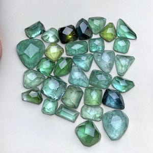 Puede incluir: Una colección de piedras preciosas facetadas sueltas en varios tonos de verde, que van desde el verde menta claro hasta el verde bosque profundo y el azul verdoso. Las piedras presentan diversas formas geométricas, incluyendo cuadrados, rectángulos, triángulos y polígonos irregulares con superficies pulidas.