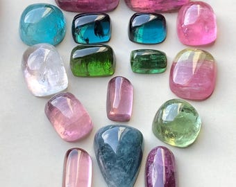May include: A collection of polished gemstones in diverse shapes and colours, including pink, blue, green, and purple. The gemstones are arranged on a light-coloured surface, showcasing their translucent qualities and natural beauty. The collection includes rectangular, oval, and teardrop-shaped stones.