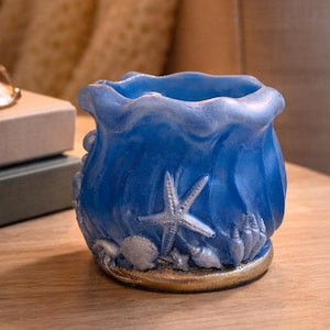 May include: A blue decorative candle holder with a wavy top and a gold base. The holder is adorned with seashells and a starfish. It sits on a wooden table, with a vase of flowers and a lit candle in the background.
