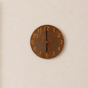 May include: A circular wall clock with a weathered, rusted brown metal finish and raised gold-toned numerals from one to twelve. The clock features two black hour and minute hands and a thin red second hand, mounted on a plain off-white wall.