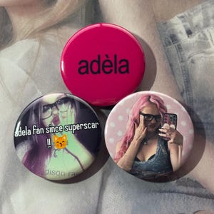 May include: Three circular pin-back buttons. The top button is solid hot pink with the black text adèla. The bottom left button features a person with glasses and the text adela fan since superscar with a cat emoji. The bottom right button shows a person with pink hair and glasses taking a selfie.
