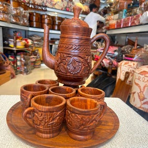 Peut inclure: Un service à thé en bois brun foncé composé d'une théière avec couvercle et anse, et de six tasses assorties ornées de motifs floraux sculptés. L'ensemble est disposé sur un plateau de service rond en bois, révélant le grain du bois.