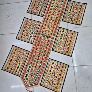 May include: A rectangular table runner and six matching rectangular placemats featuring intricate embroidery in red, green, and gold thread with small circular mirrors embedded throughout. The runner has a pointed end with a red tassel.