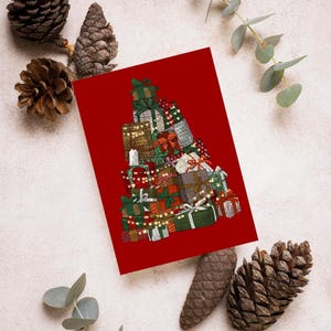 Puede incluir: Una tarjeta roja de felicitación con una ilustración de regalos de Navidad apilados, decorados con lazos verdes y luces de cadena. La tarjeta está rodeada de piñas y eucalipto, creando un ambiente festivo.