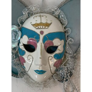 May include: A white ceramic mask with a gold crown, blue and pink accents, and silver glitter details. The mask has black eye holes and a blue painted mouth. It is decorated with lace and a blue flower.