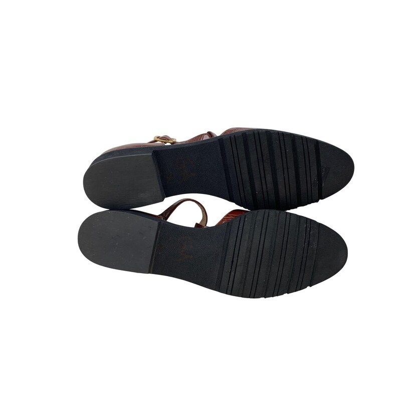 May include: Pair of brown leather T-strap sandals with a textured finish. The sandals have a small heel and a buckle closure. The soles are black with a ridged pattern for traction. The shoes are displayed against a white background.