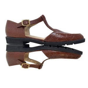 May include: Brown leather T-strap sandals with a textured, crocodile-like pattern. The sandals have a buckle closure and a black sole. The shoes are displayed on a reflective surface.