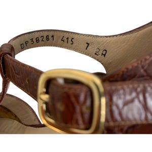 May include: Close-up of a brown leather sandal with a gold-colored buckle. The sandal has a strap with the numbers "DP38281 415 7 2A" printed on the inside. The leather has a textured pattern.