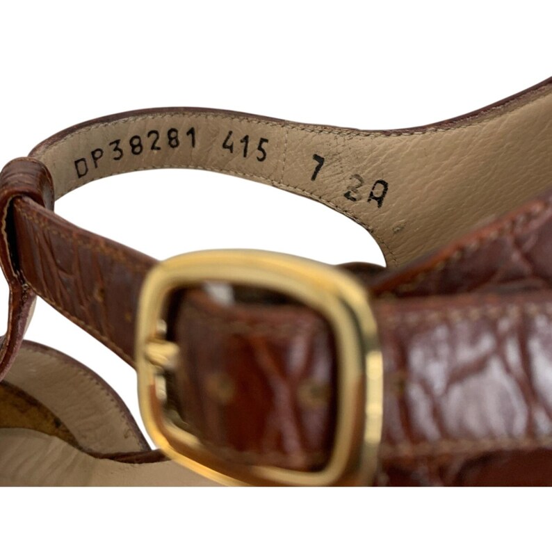 May include: Close-up of a brown leather sandal with a gold-colored buckle. The sandal has a strap with the numbers "DP38281 415 7 2A" printed on the inside. The leather has a textured pattern.