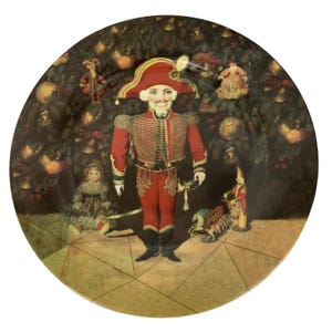 May include: A decorative plate featuring a Nutcracker soldier in a red uniform and hat, standing in front of a Christmas tree. The plate has a gold rim and a detailed illustration with a vintage aesthetic.