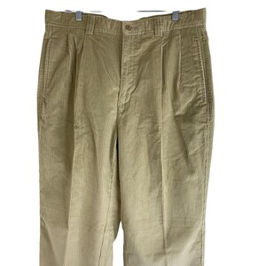 May include: Tan corduroy pants with a button closure, belt loops, and pleated front. The pants are hanging on a silver hanger. The corduroy fabric has vertical ridges.