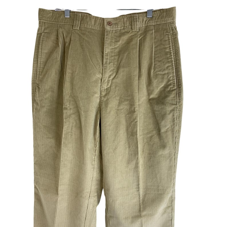 May include: Tan corduroy pants with a button closure, belt loops, and pleated front. The pants are hanging on a silver hanger. The corduroy fabric has vertical ridges.