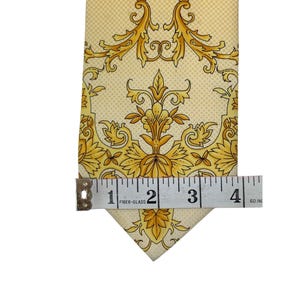 May include: A light yellow tie with a repeating gold floral pattern. The tie is measured with a tape measure, showing a width of approximately 4 inches. The tie has a pointed end.