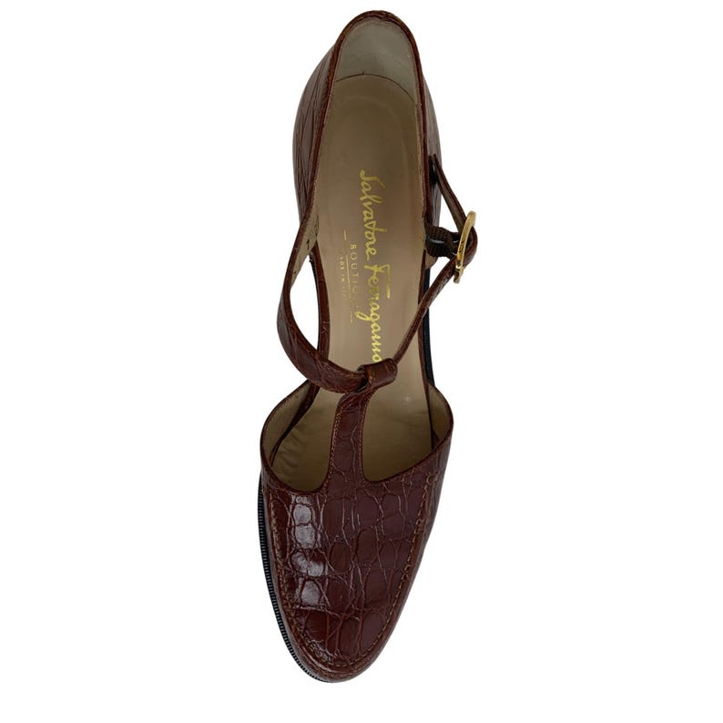 May include: A brown, high-heeled shoe with a T-strap design. The shoe features a textured, crocodile-like pattern and a gold-colored buckle. The brand name "Salvatore Ferragamo" is visible on the insole.