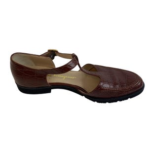 May include: Brown leather t-strap sandal with a textured, crocodile-like pattern. Features a small heel, a buckle closure, and the brand name "Salvatore Ferragamo" embossed on the side. The sole is black.
