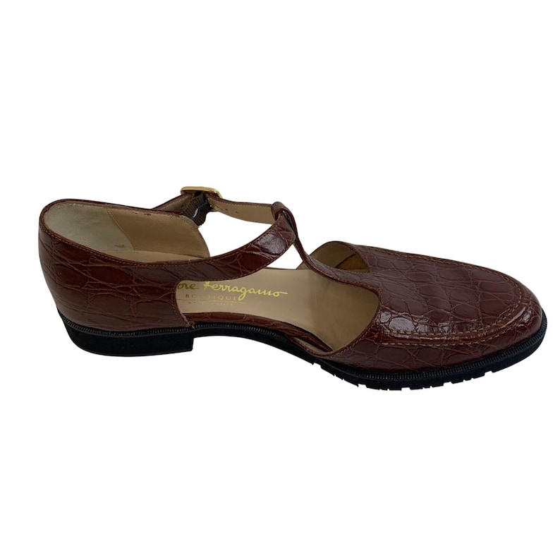 May include: Brown leather t-strap sandal with a textured, crocodile-like pattern. Features a small heel, a buckle closure, and the brand name "Salvatore Ferragamo" embossed on the side. The sole is black.