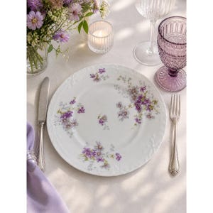 May include: A white porcelain plate with a floral design featuring purple and lavender flowers. The plate is part of a table setting, with a knife, fork, and a purple napkin. A glass of water and a candle are also present.