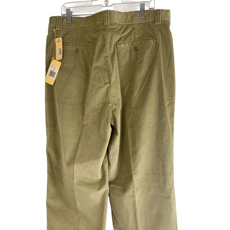May include: Back view of olive green corduroy pants hanging on a white hanger. The pants have a classic design with a button closure and pockets. A price tag is attached to the pants. The corduroy fabric has vertical ridges.