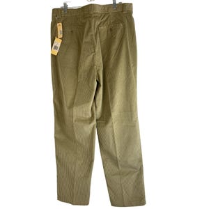 May include: Khaki-colored corduroy pants with a straight leg. The pants have a button and zipper closure, belt loops, and two back pockets. A price tag hangs from the waistband. The pants are hanging on a metal hanger.