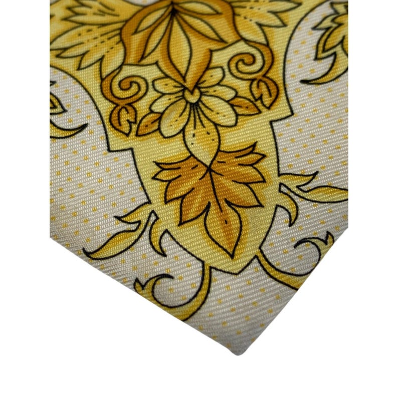 May include: Close-up of a silk tie with a repeating floral pattern in shades of yellow and gold. The tie has a cream-colored background with small, evenly spaced dots. The design features stylized flowers and leaves.