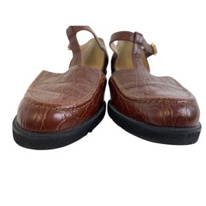 May include: Pair of brown leather T-strap shoes with a textured, crocodile-like pattern. The shoes have a rounded toe, a black sole, and a small gold buckle on the strap. The shoes are a size 8.