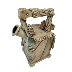 May include: A beige ceramic teapot with a unique architectural design. The teapot features a spout, handle, and a decorative roof-like structure. It has red and blue accents and a textured surface, suggesting a handcrafted appearance.